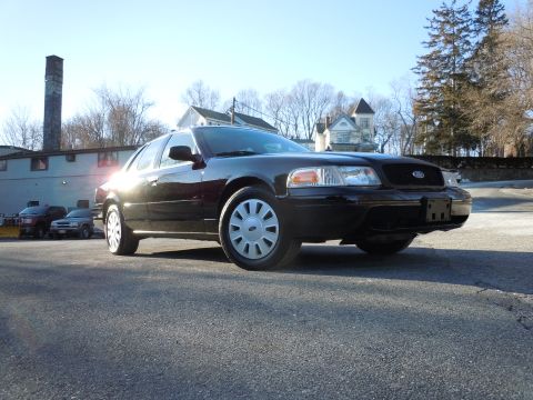 2001 Ford Crown Victoria - A black 2001 Ford Crown Victoria is parked on an asphalt surface in front of a building with a tall brick chimney. The car is viewed from a…
