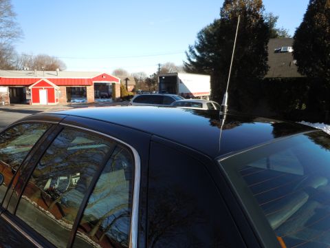 2001 Ford Crown Victoria - The glossy black roof and rear window of a sedan are shown, reflecting the surrounding trees and buildings. A tall, thin antenna extends fr…