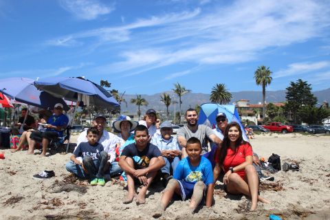 1996 Ford Bronco - A group of people are posing for a photo on a sandy beach with the ocean and mountains in the background. Two blue umbrellas are set up beh…