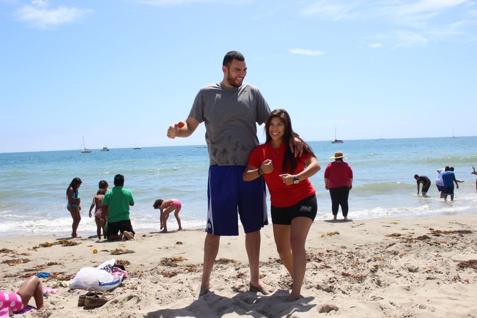 A man and woman posing on a beach with the ocean and sailboats in the background.