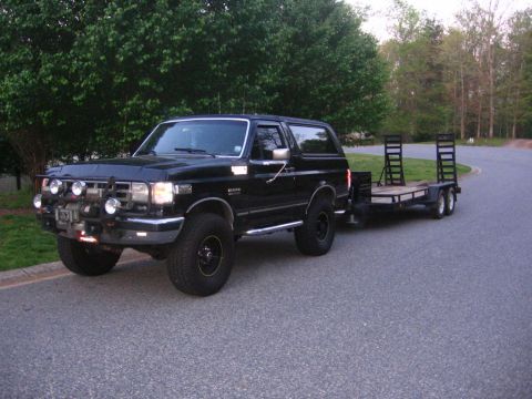 1988 Ford Bronco - A black 1980s Ford Bronco is parked on a paved road, attached to a black tandem axle trailer with ramps. The Bronco has a winch and multipl…