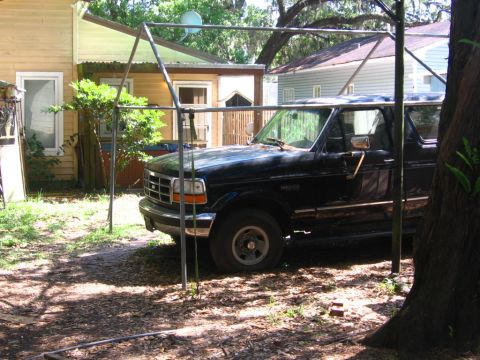 1988 Ford Bronco - A black Ford Bronco is parked under a metal frame structure in a yard with trees and houses in the background. The vehicle is viewed from t…