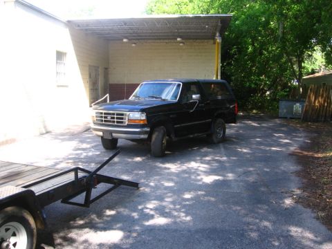 1988 Ford Bronco - A black Ford Bronco is parked under a covered carport next to a building. A trailer is partially visible in the foreground.