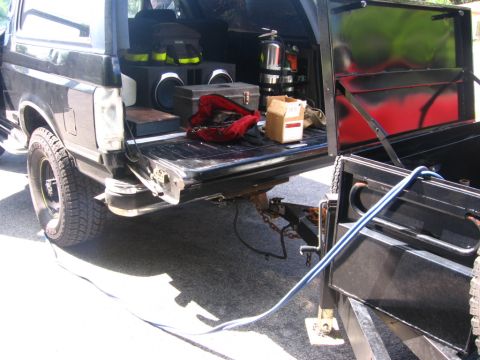 1988 Ford Bronco - The rear of a black Ford Bronco is shown with its tailgate down, revealing a large speaker box and various items. A trailer is attached to…