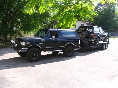 1988 Ford Bronco - A black Ford Bronco is parked on asphalt, towing a trailer with another black Ford Bronco on it. The front Bronco has aftermarket lights an…