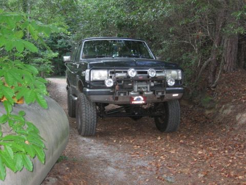 1988 Ford Bronco - A dark-colored, lifted Ford Bronco is parked on a dirt road surrounded by trees. The front of the vehicle features a brush guard with multi…