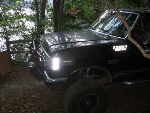1988 Ford Bronco - The front passenger side of a black Ford Bronco is visible, parked on a dirt path next to a wooden fence and trees. The vehicle features a…