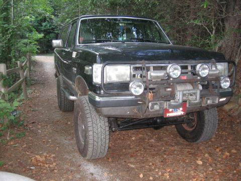 1988 Ford Bronco - A black Ford Bronco is parked on a dirt path surrounded by trees. The front of the SUV features a winch and multiple auxiliary lights mount…