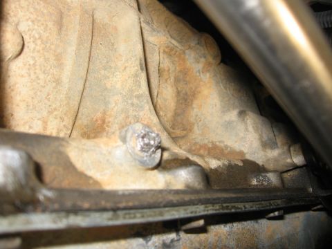 1988 Ford Bronco - A close-up view of a heavily rusted bolt on a vehicle's transmission or transfer case housing. The bolt head is partially obscured by what…