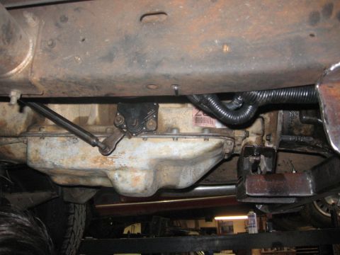 1988 Ford Bronco - The underside of a vehicle shows the transmission pan, a linkage mechanism, and a black corrugated hose attached to the frame.