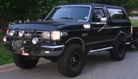 1988 Ford Bronco - A black two-door Ford Bronco with a brush guard, winch, and auxiliary lights is parked on a paved surface with trees in the background.