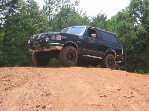 1988 Ford Bronco - A dark-colored, 1980s Ford Bronco is angled on a dirt mound with trees in the background. The SUV is shown from a 3/4 front view, highlight…