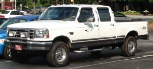 1988 Ford Bronco - A white four-door pickup truck is parked in a lot. The truck has chrome accents on the front grille and wheels, and a black stripe along th…