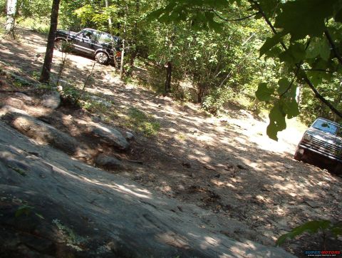 1988 Ford Bronco - Two dark-colored SUVs are seen off-roading on a steep, rocky, and wooded trail. The lead SUV is a black Ford Bronco, and the second SUV is…