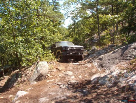 1988 Ford Bronco - A dark-colored 1988 Ford Bronco is navigating a rocky, wooded trail. The vehicle is positioned on a dirt path surrounded by trees and large…