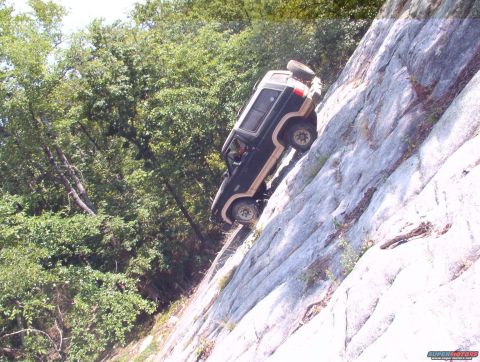 1988 Ford Bronco - An off-road vehicle navigates a challenging ascent up a steep, rocky mountainside, with a spare tire visible on its roof.