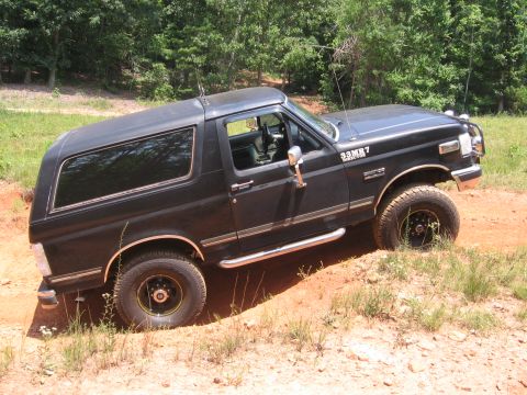 1988 Ford Bronco - A black Ford Bronco is shown driving off-road on a dirt path with trees in the background. The vehicle is angled to the right, showing its…