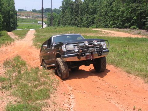 1988 Ford Bronco - A black Ford Bronco is driving off-road on a dirt path with grassy areas on either side. The vehicle has a brush guard with auxiliary light…