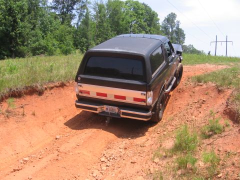 1988 Ford Bronco - A black Ford Bronco is shown from a rear three-quarter angle, driving on a dirt road with red clay. The vehicle has a chrome bumper and a r…