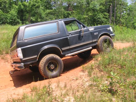 1988 Ford Bronco - A black 1980s Ford Bronco is shown from a 3/4 rear angle, driving on a dirt path surrounded by trees and tall grass. The vehicle has chrome…