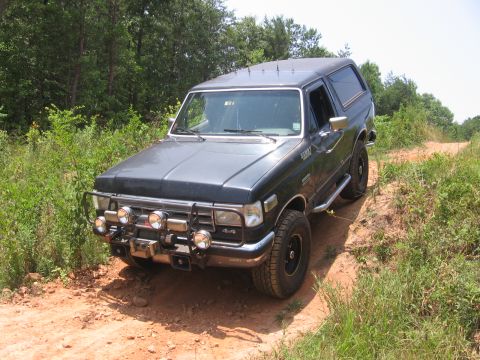 1988 Ford Bronco - A dark gray Ford Bronco is parked on a dirt path surrounded by lush green vegetation and trees. The vehicle has a brush guard with multiple…