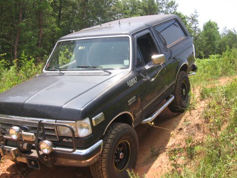 1988 Ford Bronco - A black Ford Bronco is parked on a dirt path surrounded by trees and vegetation. The vehicle has a brush guard with two large fog lights mo…