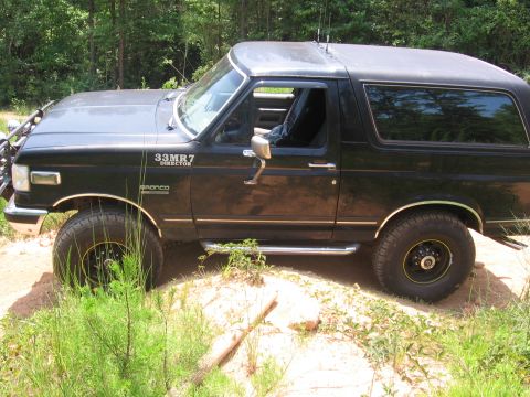 1988 Ford Bronco - A black Ford Bronco is parked on a dirt path surrounded by trees. The vehicle has off-road tires with yellow accents on the rims and a chro…