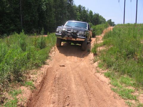1988 Ford Bronco - A black Ford Bronco is driving up a steep, rutted dirt road surrounded by green foliage and trees. The vehicle is angled towards the camera…