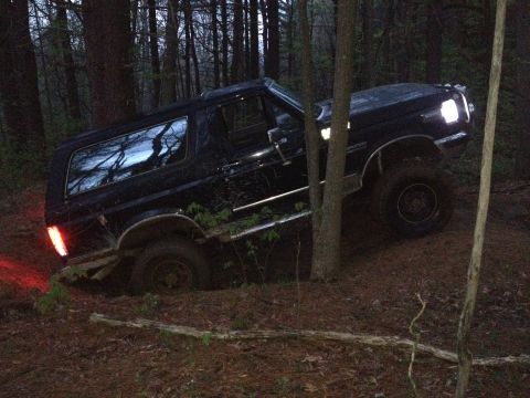 1988 Ford Bronco - This black SUV navigates a wooded trail, its front lights illuminated against the dim forest surroundings.