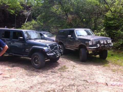 1988 Ford Bronco - Two off-road vehicles, a 1988 Ford Bronco and a Jeep Wrangler, are parked on a dirt path in a wooded area.