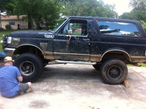 1988 Ford Bronco - A person works on a lifted, muddy Ford Bronco parked on a concrete surface.