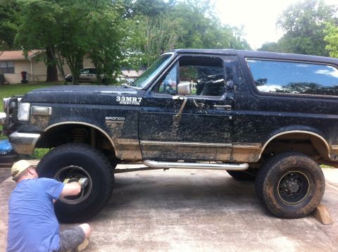 1988 Ford Bronco - A person works on the front wheel of a muddy black Ford Bronco, seen from the side.
