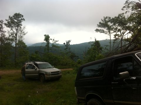 1988 Ford Bronco - A black Ford Bronco is parked on a dirt road with a silver SUV in the background, surrounded by trees and hills.