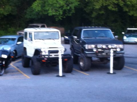 1988 Ford Bronco - Two modified off-road vehicles are parked in a lot, one black pickup and one white SUV.