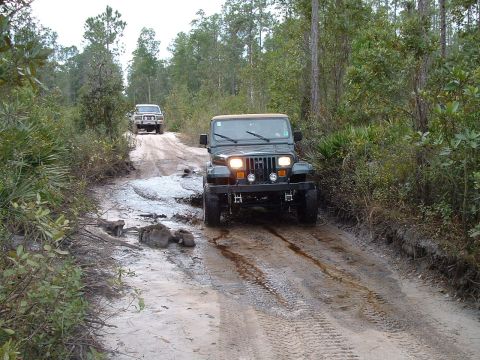 1995 Ford Bronco - Two SUVs navigate a challenging muddy path through a wooded area.