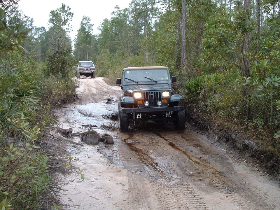 Green Jeep Wrangler driving on a muddy dirt road, front view. Another SUV is visible in the background
