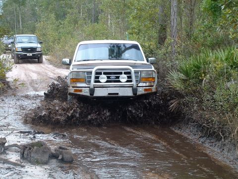 1995 Ford Bronco - A white 1995 Ford Bronco navigates a muddy trail, kicking up a large splash as it drives through a water obstacle.