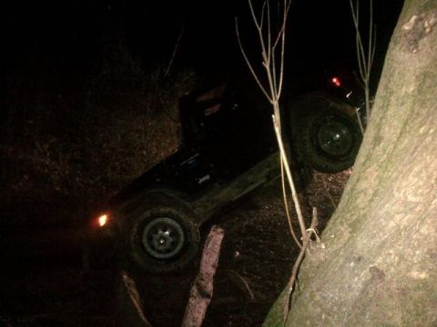 1988 Ford Bronco - A black Jeep is seen tilted on its side, partially obscured by trees and darkness.