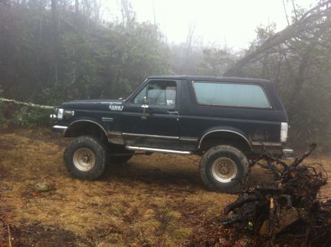 1988 Ford Bronco - This 1988 Ford Bronco is shown off-road in a foggy wooded area, equipped with large tires for rough terrain.