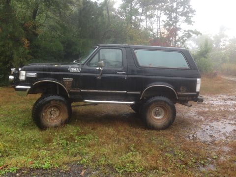 1988 Ford Bronco - Side view of a black Ford Bronco SUV with aftermarket wheels and tires, parked outdoors on a damp day.