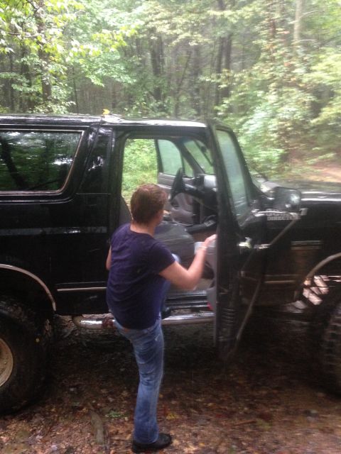 1988 Ford Bronco - A person enters a black SUV parked on a muddy trail surrounded by trees.