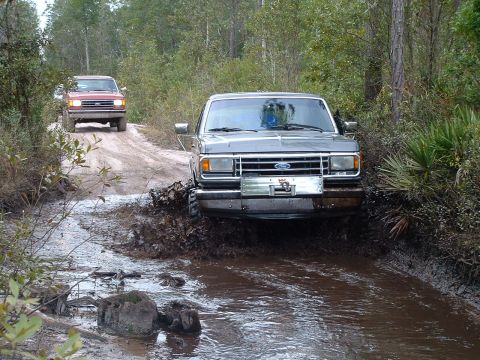 1995 Ford Bronco - Two Ford Broncos navigate a muddy trail through a wooded area.