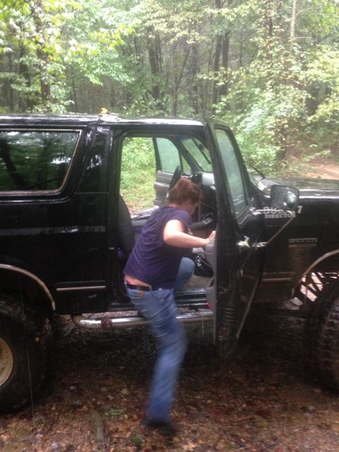 1988 Ford Bronco - A person climbs into a black 1988 Ford Bronco parked off-road in a wooded area.