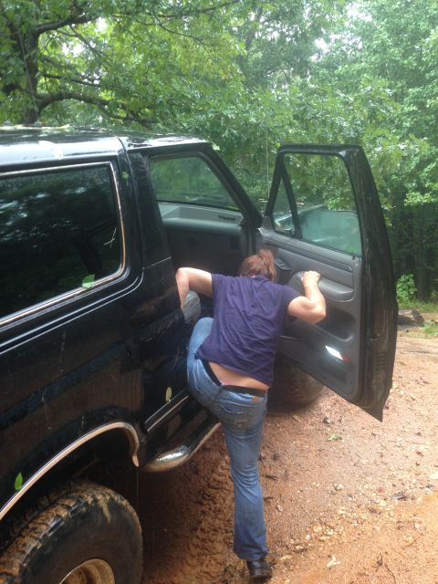 1988 Ford Bronco - A person climbs into a black SUV stuck in mud, surrounded by trees.