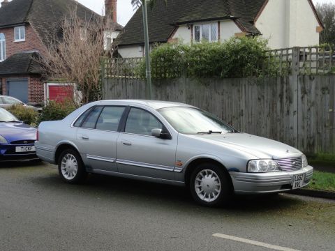 1999 Ford Fairlane - A silver sedan is parked on a residential street next to a blue car.