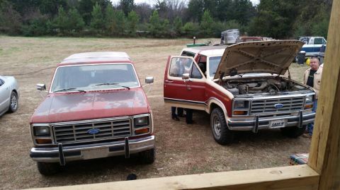 1986 Ford Bronco - Two Ford Broncos are parked in a grassy area with trees in the background. One has its hood open.