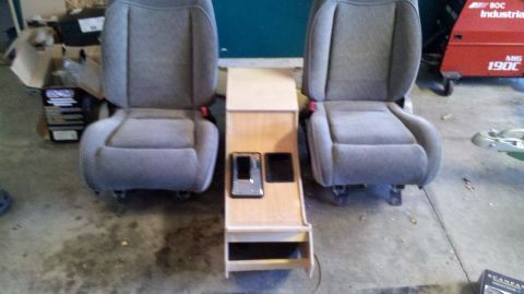 1982 Ford Bronco - Two gray fabric car seats are positioned facing forward with a wooden console in between them. The console has two compartments on top, one…