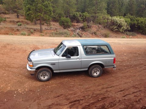 1996 Ford Bronco - This silver Ford Bronco with a green top is parked on a dirt road with trees in the background.