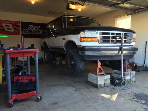 1994 Ford Bronco - A two-tone Ford Bronco is raised on jack stands inside a garage, undergoing maintenance.