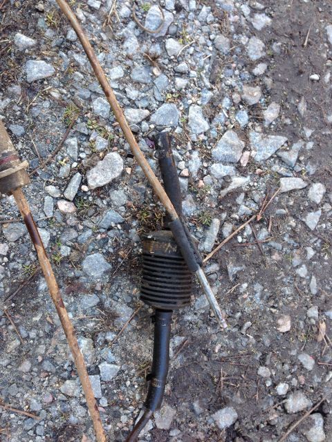 1979 Ford Bronco - Two rusty metal rods and a black ribbed component with a metal end lie on a gravel and dirt surface.